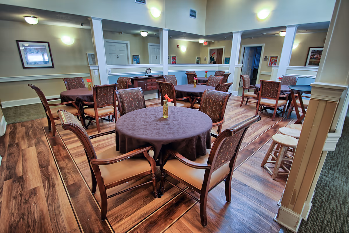A dining area with multiple round tables covered with dark tablecloths, each surrounded by wooden chairs with cushioned seats and backs. Small vases with red flowers are placed on each table. The room has wood-patterned flooring, beige walls with white trim, and several framed pictures on the walls. There is a small wooden sideboard against one wall and a few wooden stools near a counter on the right side.