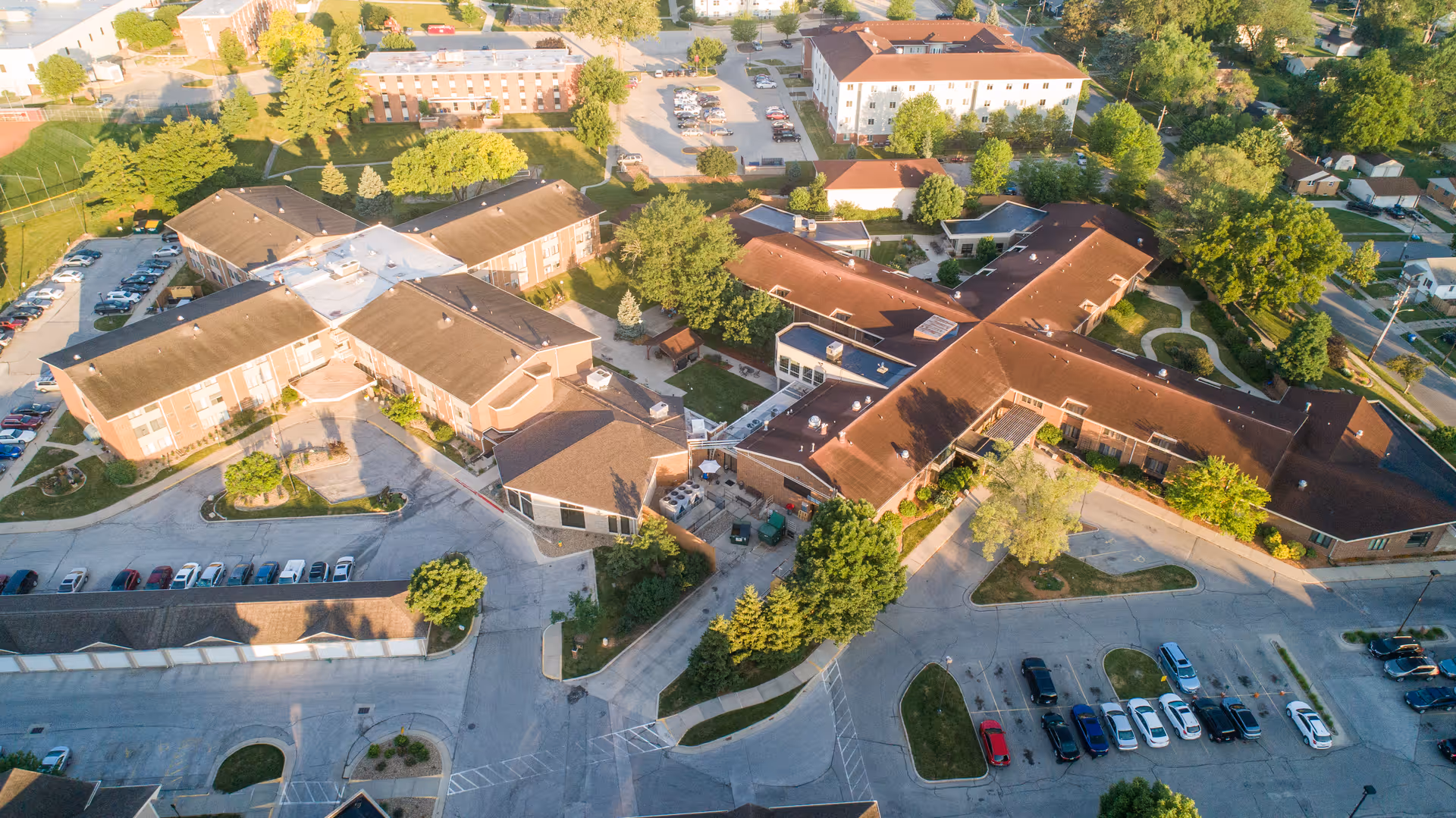 Aerial view of Luther Park Community showing multiple connected buildings with brown roofs, surrounded by trees, parking lots with cars, and walkways in a suburban area.