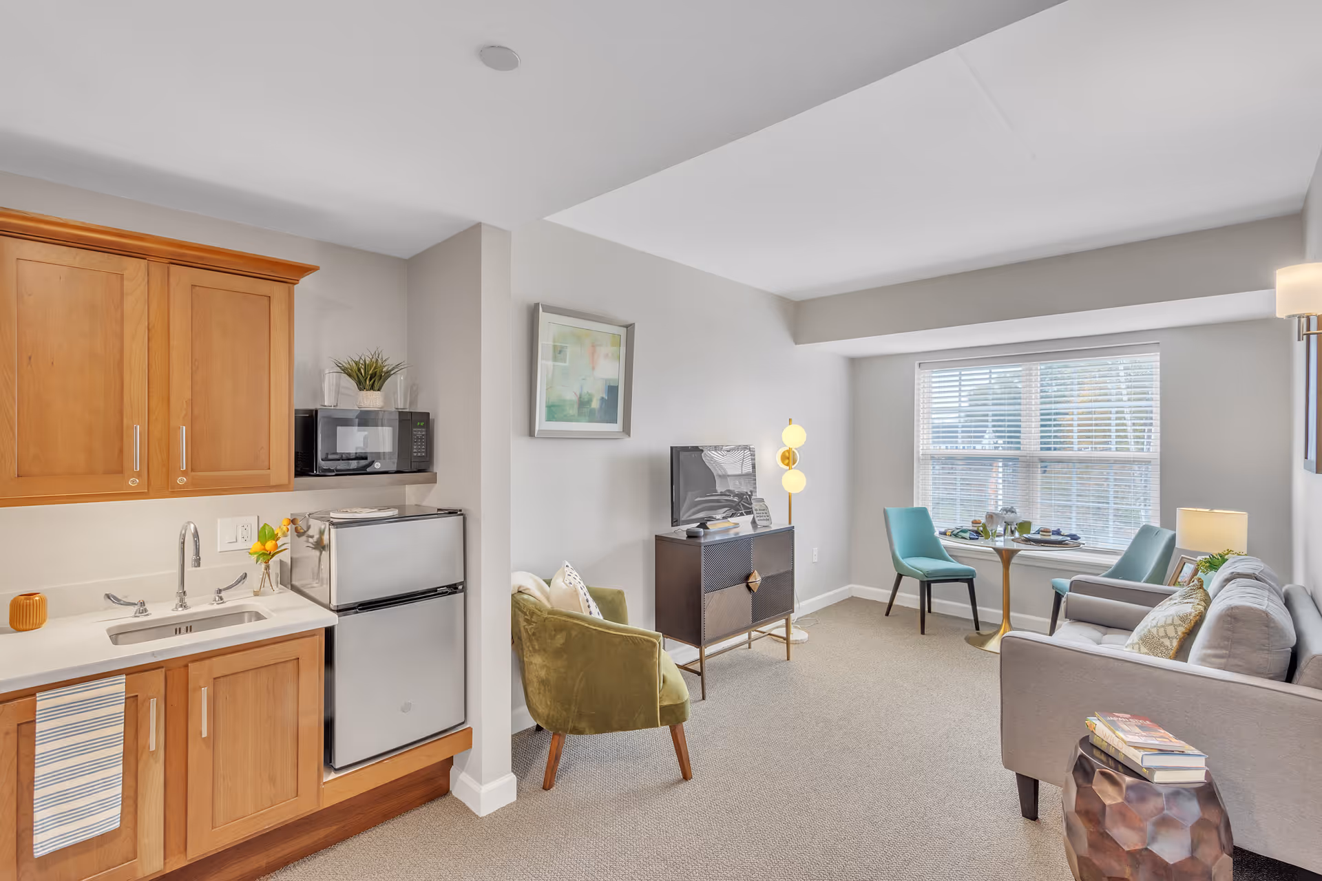 Bright, open living area with a small kitchenette on the left, seating, TV cabinet, and a dining nook by a window.