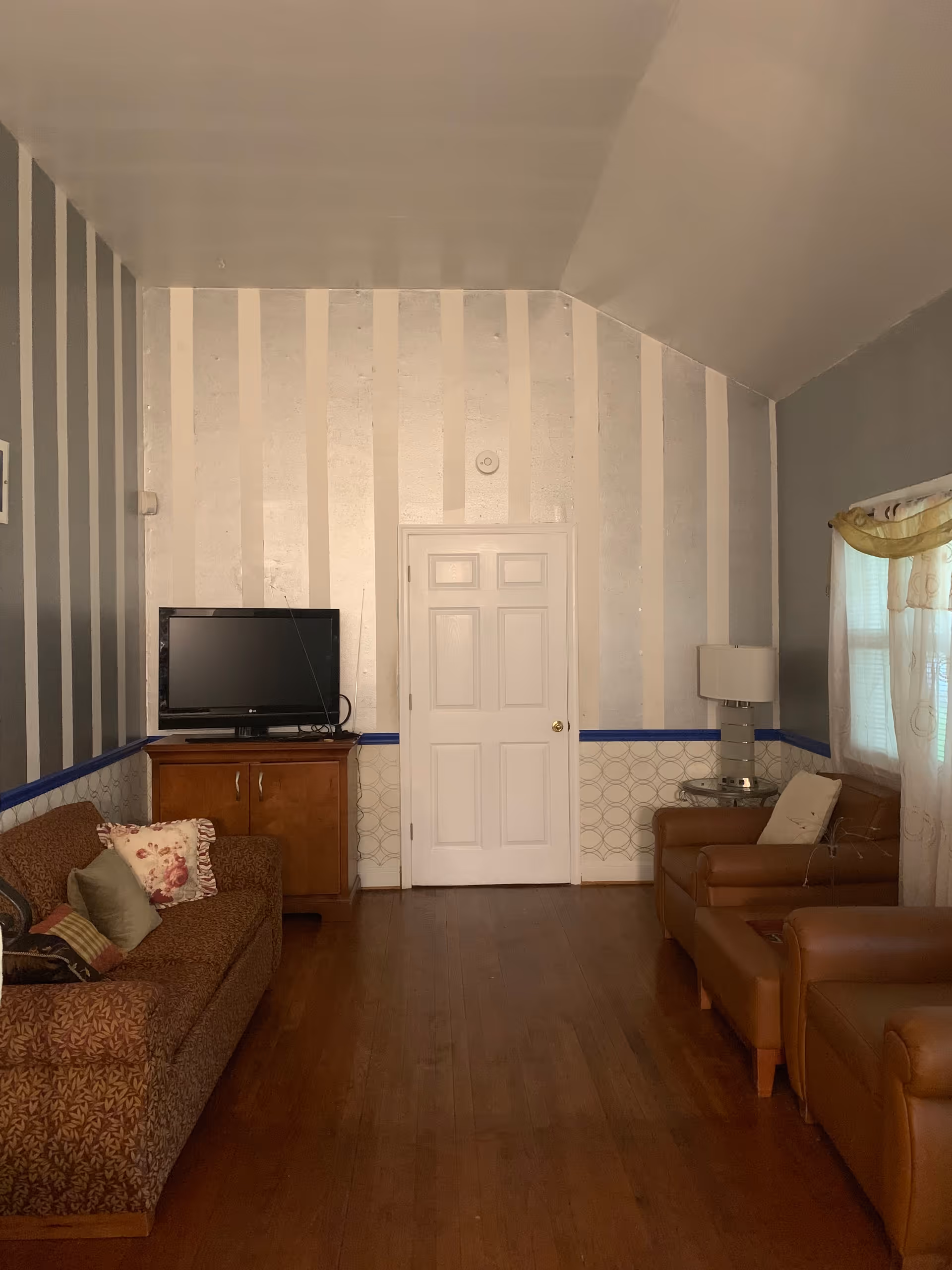 A cozy living room with striped wallpaper and wooden flooring. The room features a brown patterned sofa with decorative pillows on the left, a wooden cabinet with a flat-screen TV on top, and two brown leather armchairs on the right next to a window with sheer curtains and a yellow valance. A small round side table with a lamp is positioned between the armchairs. A white door is centered on the far wall.