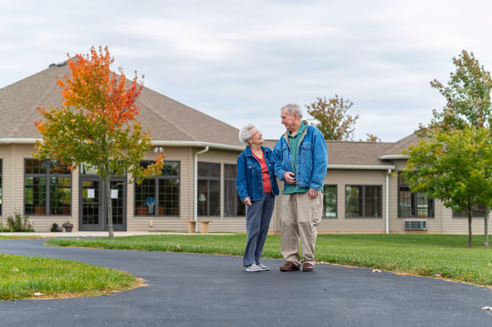 An elderly couple standing on a paved pathway outside a single-story building with large windows. The woman and man are smiling at each other, both wearing denim jackets. Trees with autumn foliage surround the area under a cloudy sky.