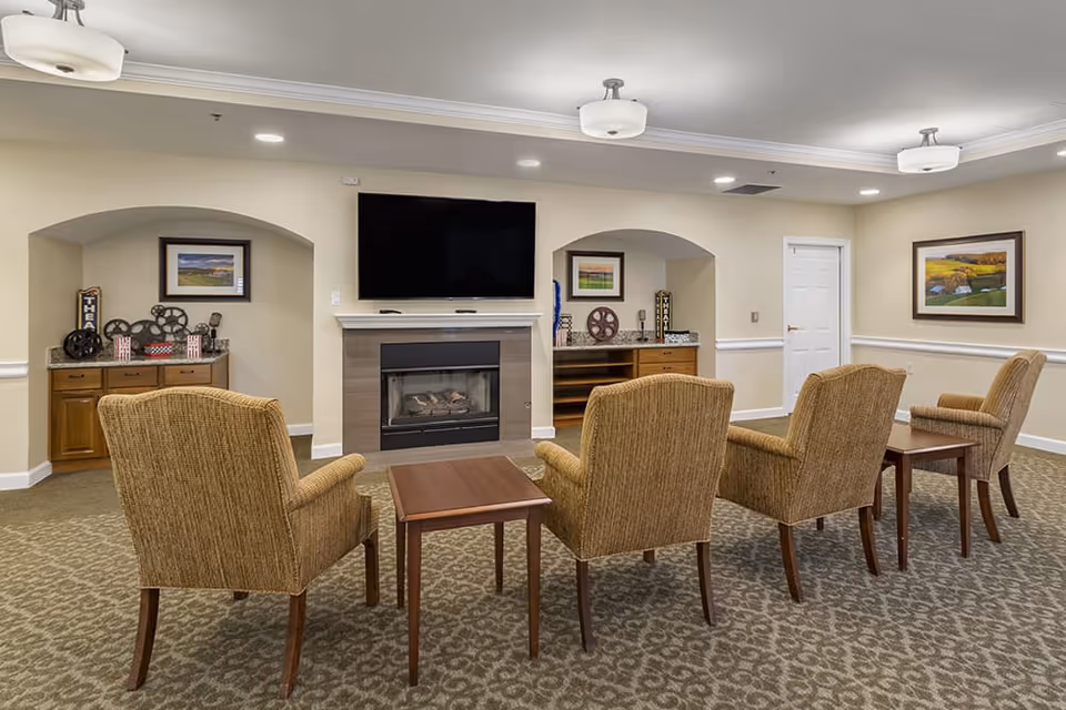 A cozy living room area with four upholstered armchairs arranged in two rows facing a wall-mounted flat-screen TV above a modern fireplace. The room has beige walls, patterned carpet, two wooden side tables, and built-in shelves with decorative items including vintage film reels and framed pictures. Ceiling lights provide soft illumination.