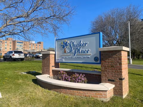 Outdoor view of a large sign for Shaker Place Rehabilitation & Nursing Center, mounted on a brick structure with a curved bench and flowers in front. The background shows a parking lot with cars, trees without leaves, and a building under a clear blue sky.