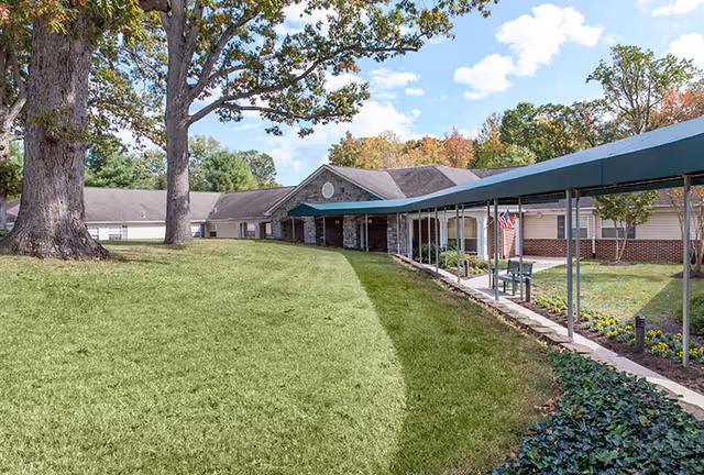 Exterior view of a senior living facility with a covered walkway leading to the entrance. The building is single-story with a mix of brick and siding, surrounded by green grass, trees, and landscaped flower beds under a partly cloudy sky.