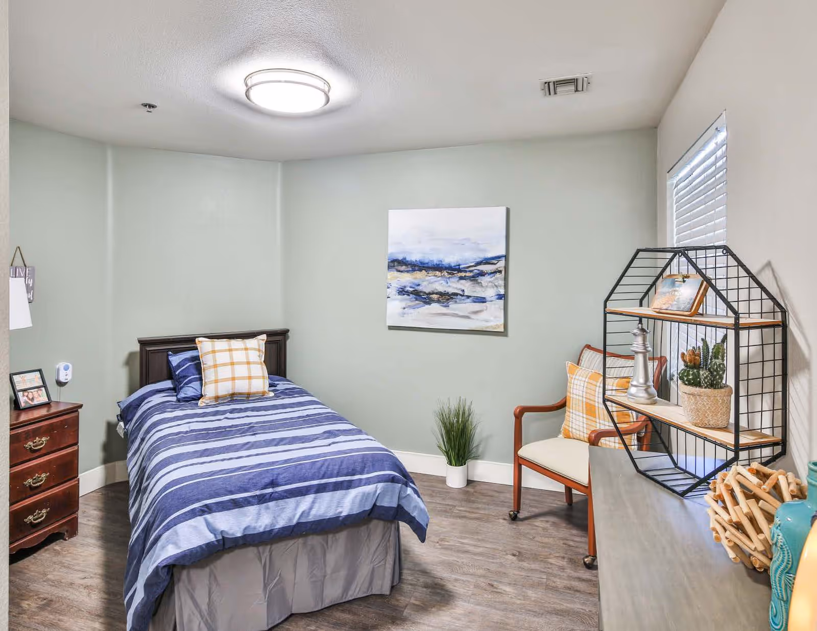 A cozy bedroom with a single bed covered in blue and white striped bedding and a plaid pillow. There is a wooden nightstand with drawers and a framed photo on it. A modern light fixture is on the ceiling. The walls are painted light green, and there is a piece of abstract art hanging above the bed. A wooden chair with a plaid cushion and a decorative shelving unit with plants and ornaments are also visible. The floor has a wood-like finish.