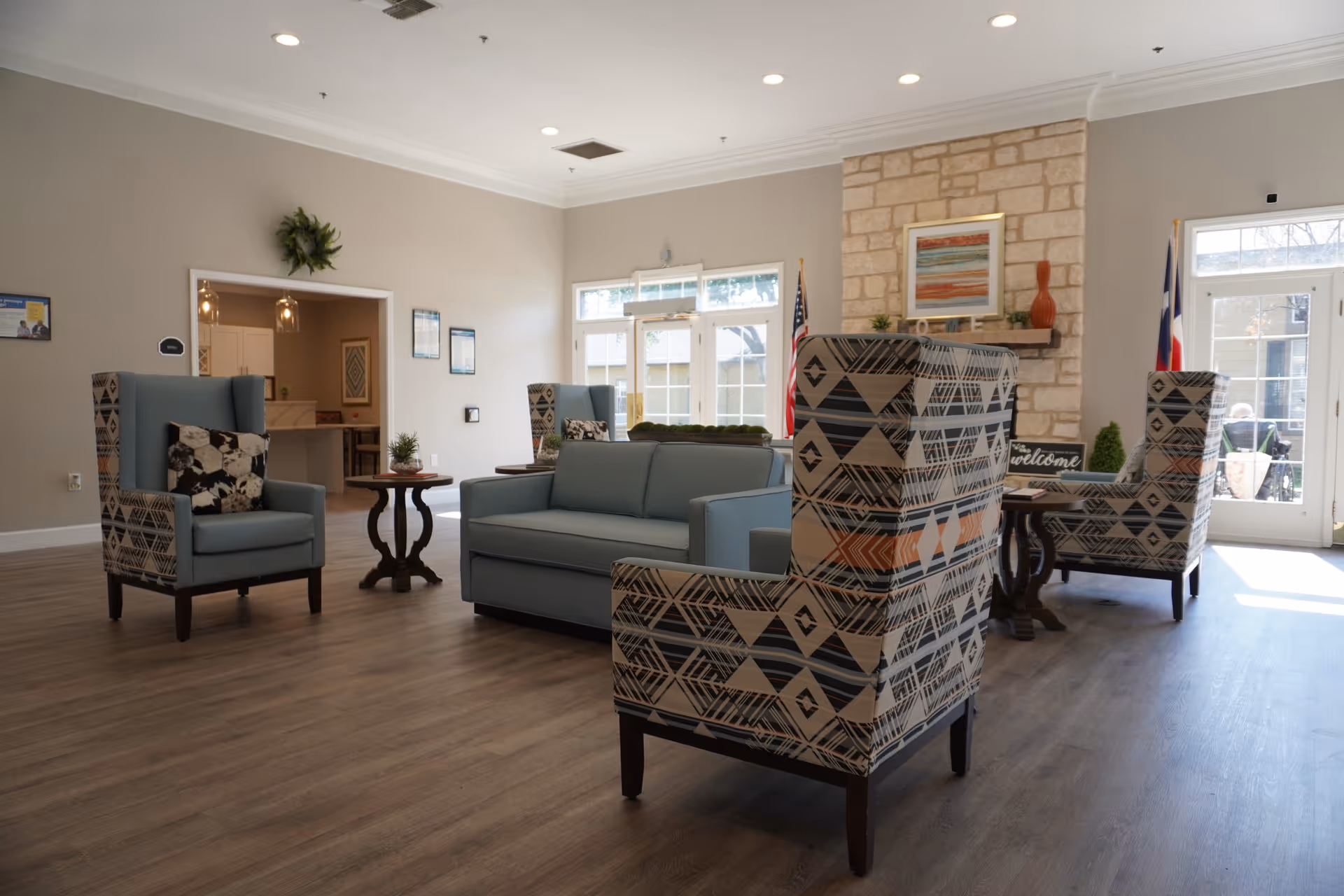 A bright and spacious living room area in a senior living facility featuring a light blue sofa, patterned armchairs, wooden side tables with small plants, a stone fireplace with a framed artwork above it, and large windows and glass doors letting in natural light. The room has wood flooring and neutral-colored walls.