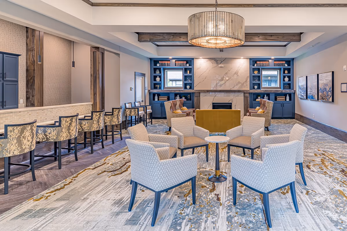 A spacious and well-lit living room area with a circular arrangement of six patterned armchairs around a small round table on a decorative carpet. In the background, there are two blue built-in bookshelves flanking a modern fireplace with a marble surround. To the left, there is a bar counter with six high chairs featuring floral upholstery. The ceiling has wooden beams and a large, round chandelier.