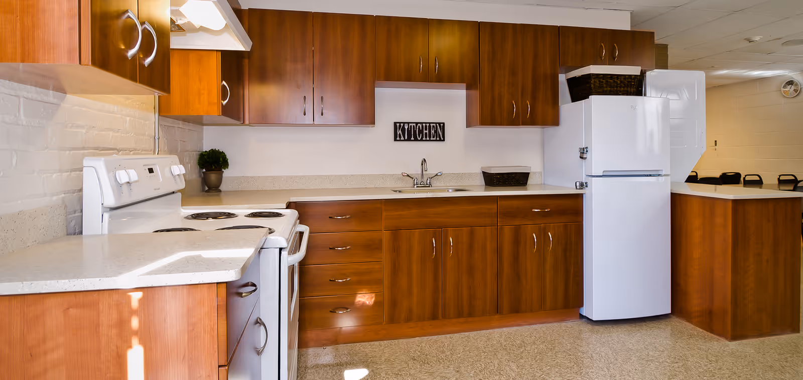 A clean and well-lit kitchen with wooden cabinets, a white stove, a white refrigerator, a countertop with a sink, and a small plant. A sign on the wall above the sink reads 'KITCHEN'.