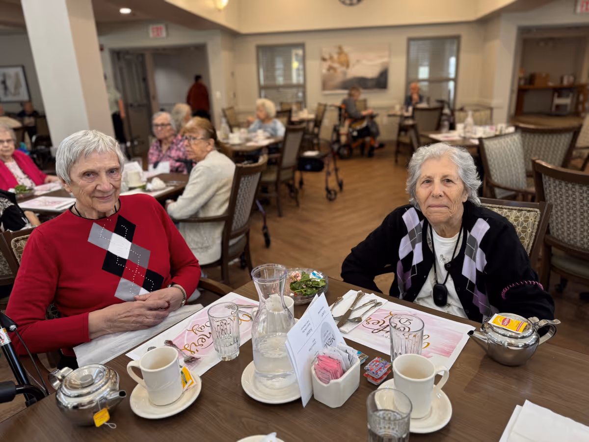 Two elderly women sitting at a dining table in a senior living facility dining room. The table is set with cups, glasses, a water pitcher, a teapot, and condiments. Other elderly residents are seated at tables in the background, and the room has a warm, inviting atmosphere with wooden floors and neutral-colored walls.