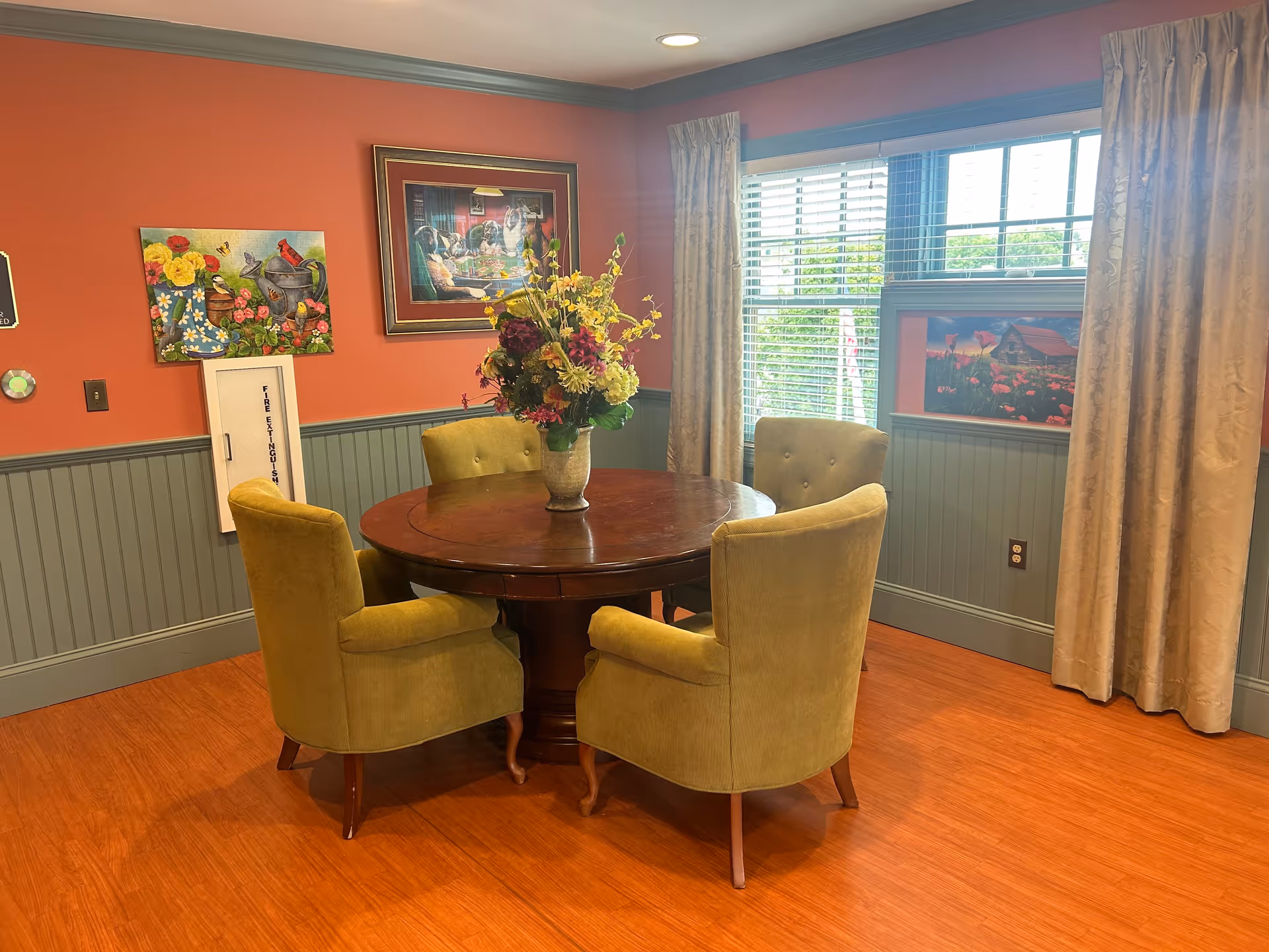 A cozy dining area with a round wooden table surrounded by four green upholstered chairs. A vase with a colorful flower arrangement sits in the center of the table. The walls are painted orange with green wainscoting, and there are three framed pictures hanging on the walls. A window with beige curtains allows natural light into the room.