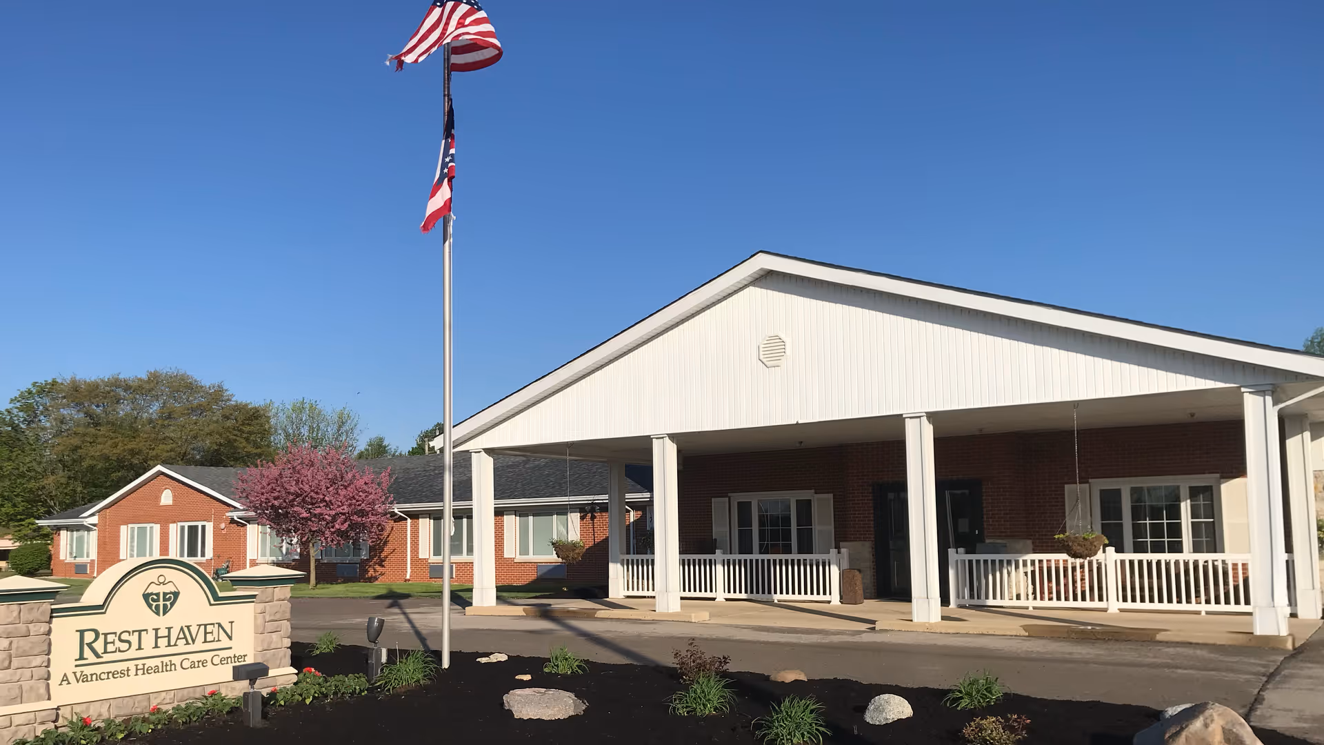 Front exterior view of Rest Haven, a Vancrest Health Care Center, showing a single-story brick building with white trim, a covered entrance supported by white columns, an American flag on a flagpole, and landscaped grounds with a flowering tree and green plants.