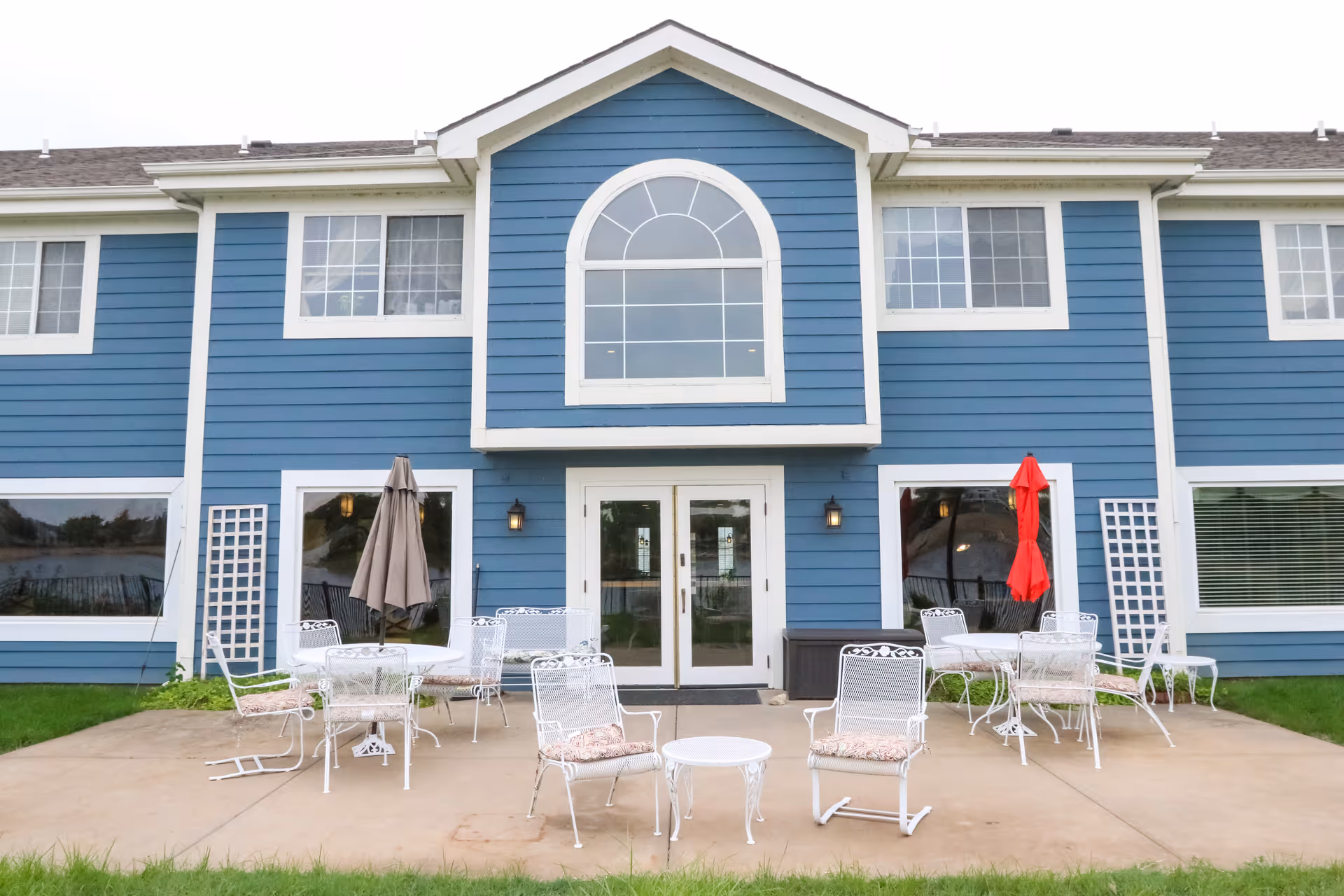 Outdoor patio area of a blue two-story building with white trim, featuring multiple white metal tables and chairs with cushions, two umbrellas (one beige and one red), and a concrete patio surrounded by green grass.