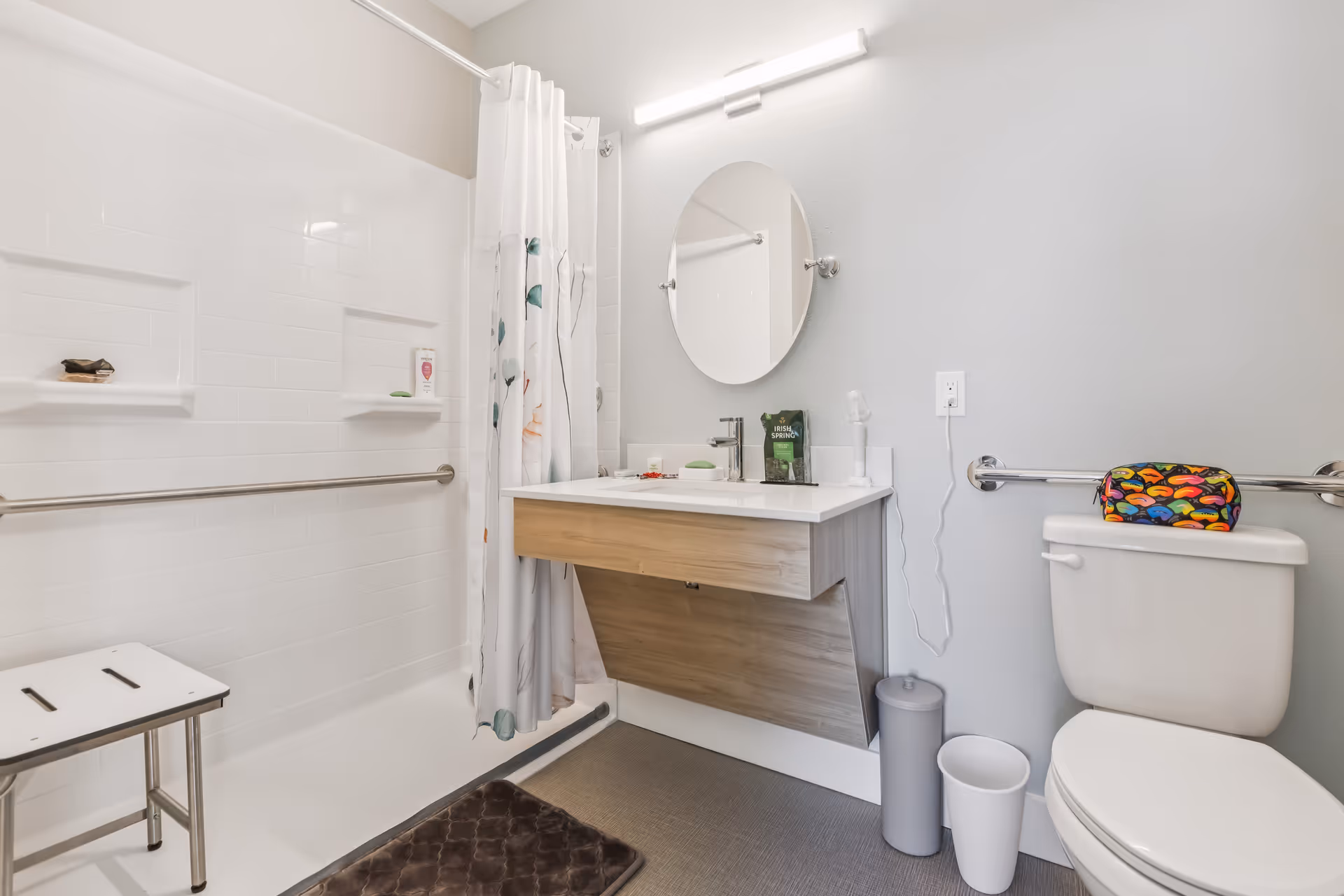 A clean and modern bathroom featuring a white tiled shower area with a shower chair and a floral shower curtain. There is a wooden floating vanity with a white countertop, a round mirror above it, and a light fixture. Next to the vanity is a white toilet with a colorful pouch on top, grab bars on the walls, and two waste bins on the floor.