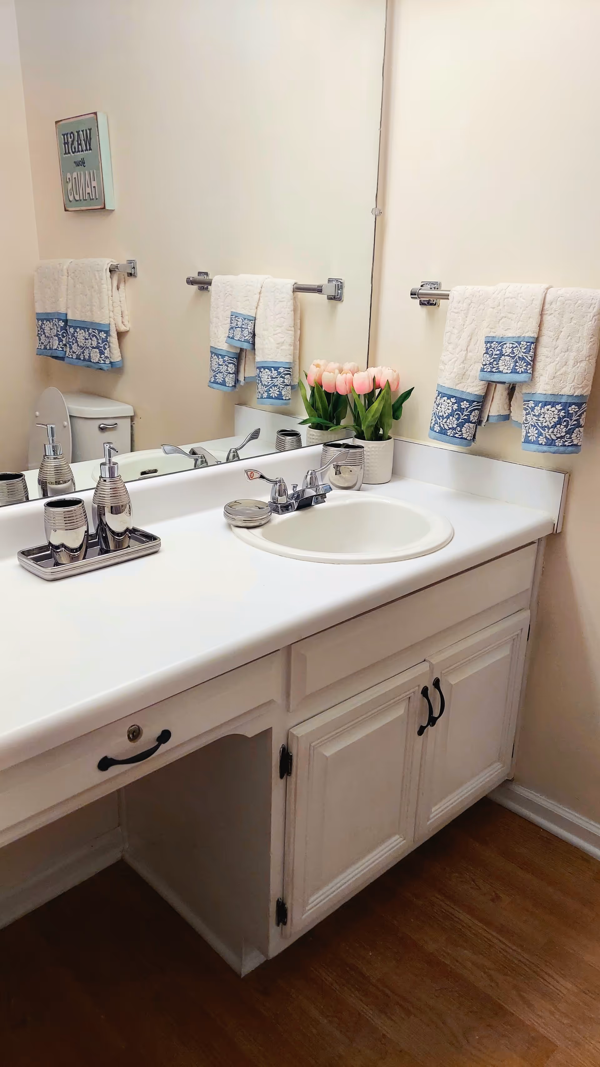 A clean bathroom vanity with a white countertop and sink, silver faucet, soap dispenser, and toothbrush holder on a tray. There are two white towels with blue floral borders hanging on a towel rack above the toilet. A small pot of pink tulips is placed near the corner of the countertop. A large mirror covers the wall behind the sink and towels. The floor is wooden.