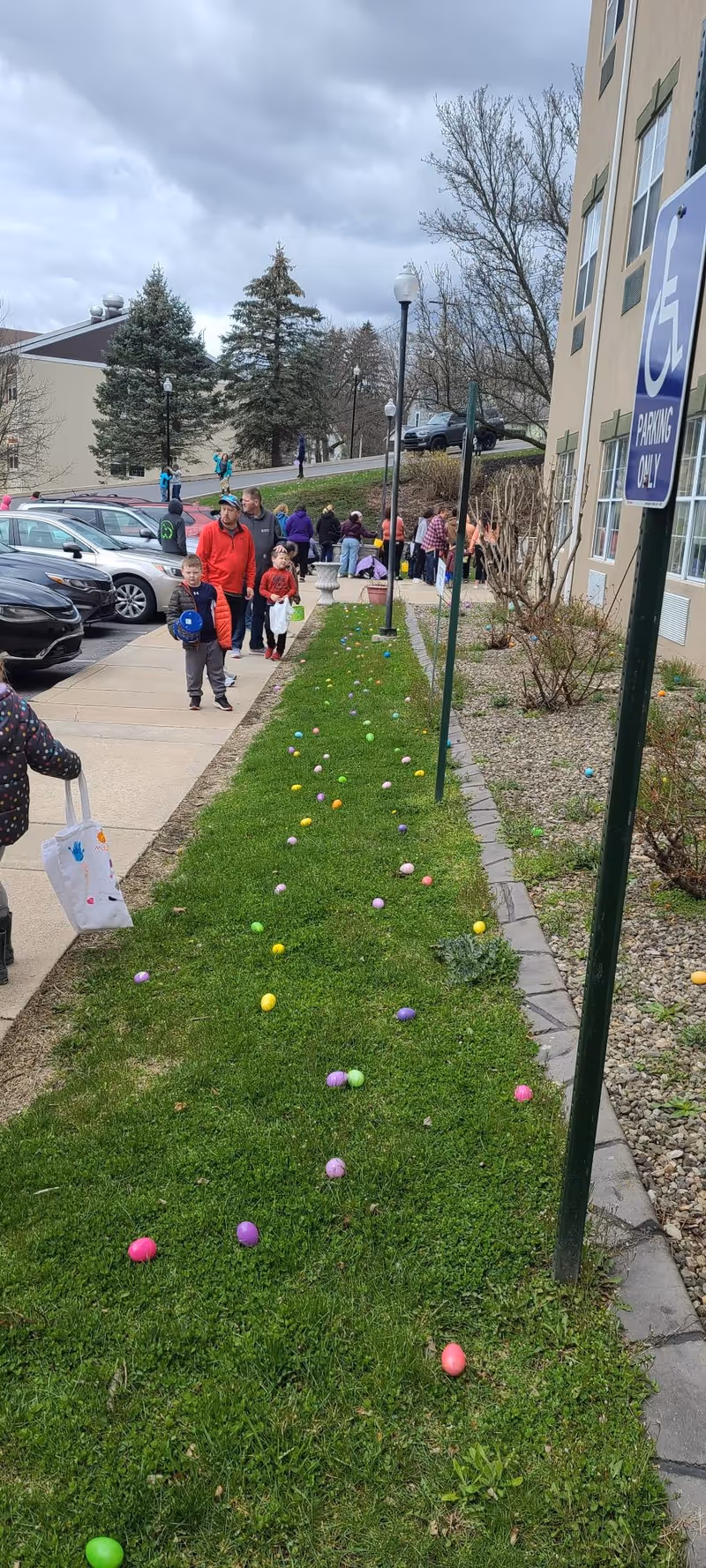 Outdoor scene at Rose View Nursing and Rehabilitation Center showing a grassy area with colorful Easter eggs scattered on the ground. Several people, including children holding bags, are walking along a sidewalk next to the grass. A building with windows and a handicapped parking sign is visible on the right side. Trees and parked cars are in the background under a cloudy sky.