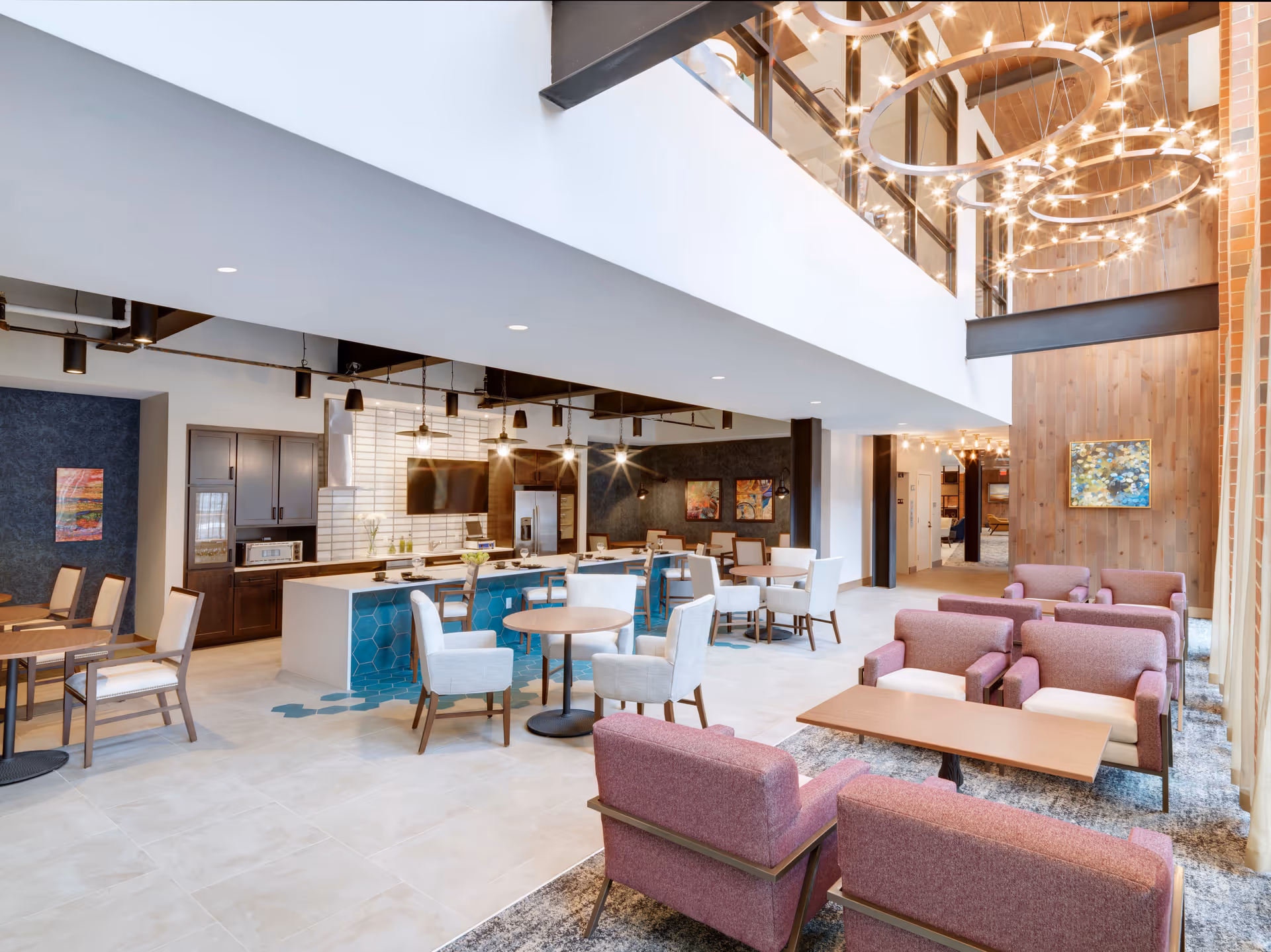A spacious and modern common area in a senior living facility featuring a seating area with purple armchairs and wooden tables on a patterned rug. The room has a high ceiling with large circular chandeliers and a wood-paneled wall with artwork. In the background, there is a kitchen area with a long island, bar stools, pendant lights, and dining tables with chairs.