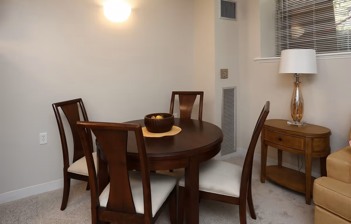 A small dining area with a round dark wooden table surrounded by four matching chairs with white cushions. On the table is a wooden bowl with fruit. To the right is a wooden side table with a decorative lamp on it, next to a beige sofa. The walls are light-colored, and there is a window with blinds above the side table.