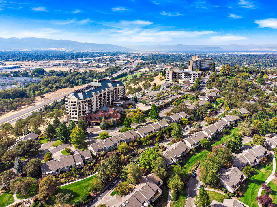 Aerial view of a large senior living complex and surrounding townhomes set among trees with roads and mountains in the distance.