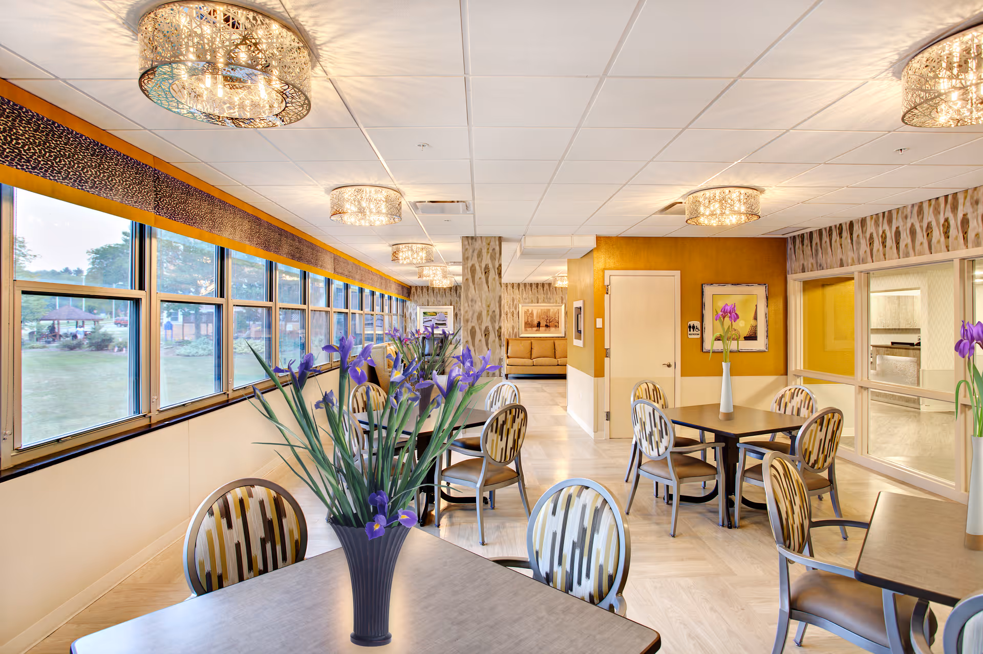 Brightly lit dining room with tables and chairs, vases of purple flowers, large windows along one wall, and decorative ceiling lights.