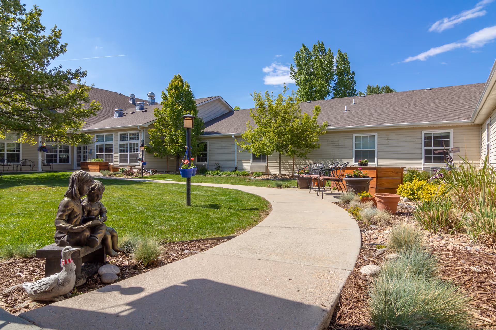 Sunny courtyard with a curved concrete path, lawn, outdoor seating, planters, and a bronze statue of two children reading in front of a single-story building.