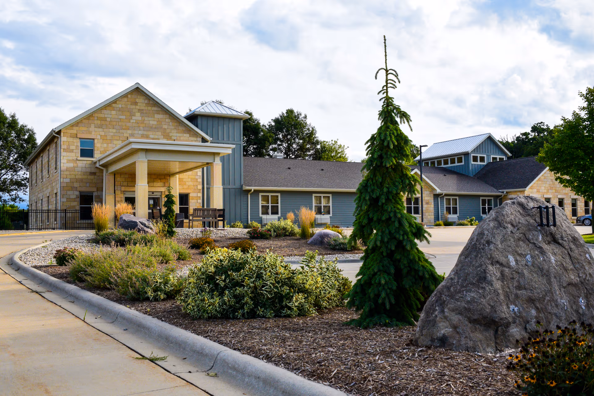 Exterior front view of a single-story memory care building with a covered entrance, landscaped roundabout, and a large rock in the foreground.