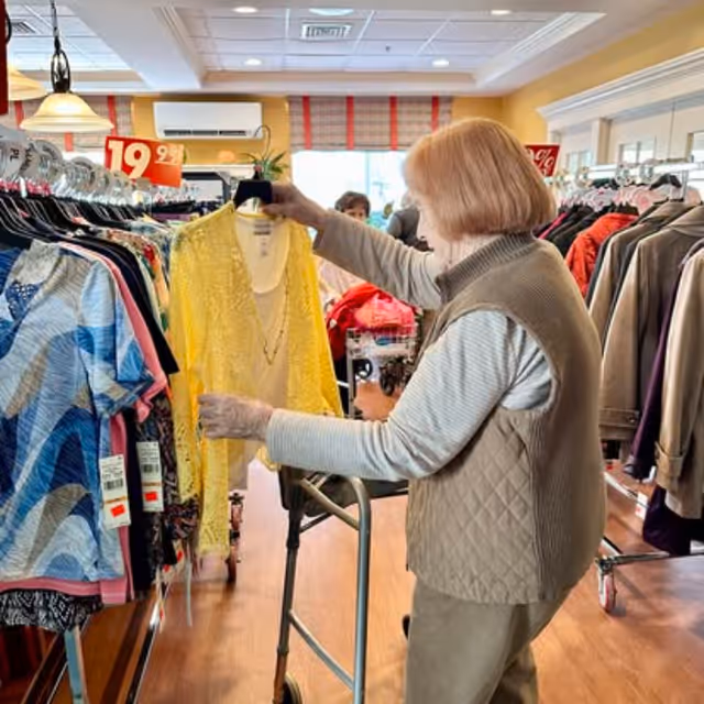 An elderly woman using a walker is shopping for clothes in a store, holding a yellow cardigan on a hanger while standing next to racks filled with various garments.