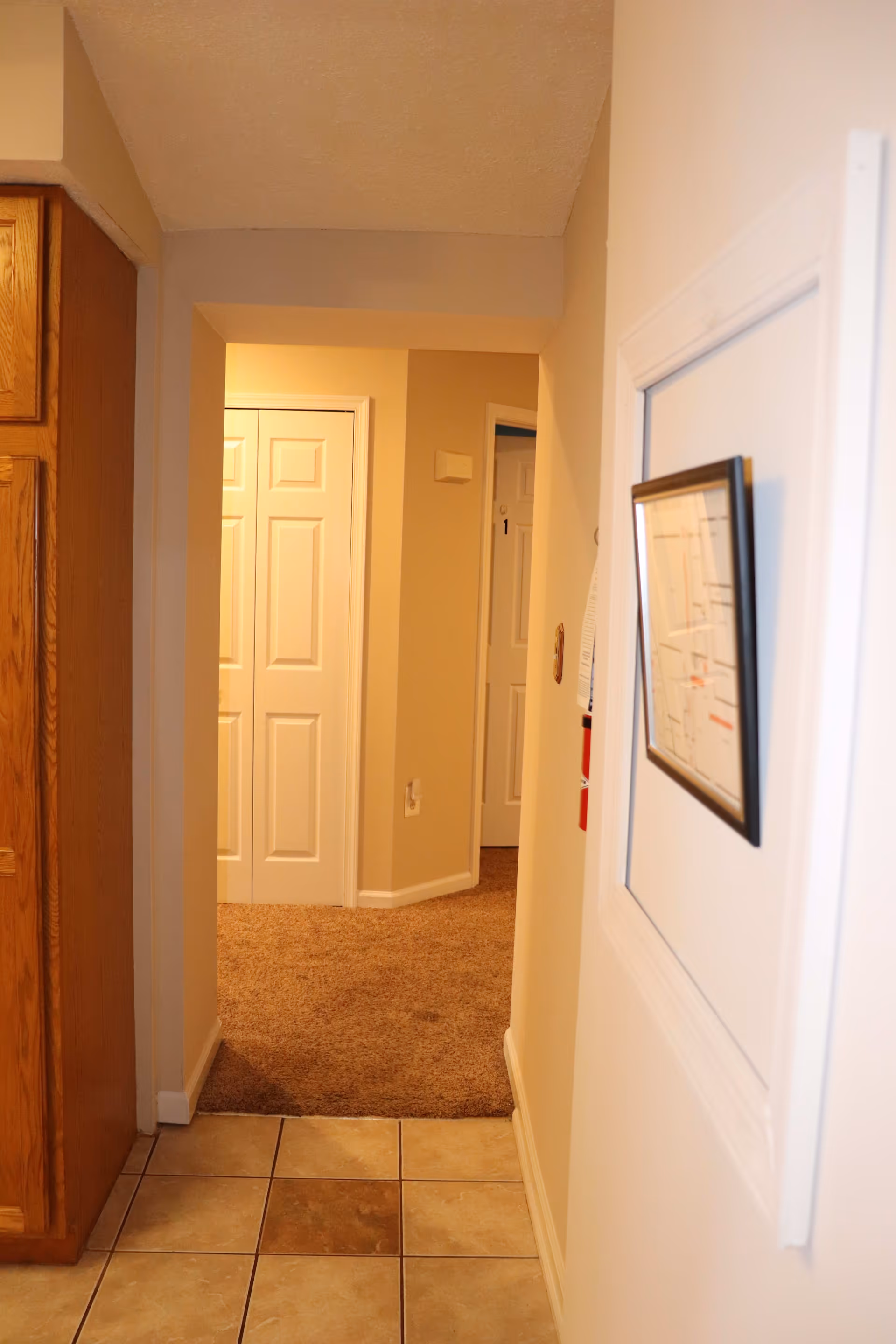 Interior view of a hallway in a residential facility with tiled flooring leading to a carpeted area. There are white double doors at the end of the hallway and a door labeled with the number 1. A framed map or diagram is mounted on the right wall, and wooden cabinetry is visible on the left side.