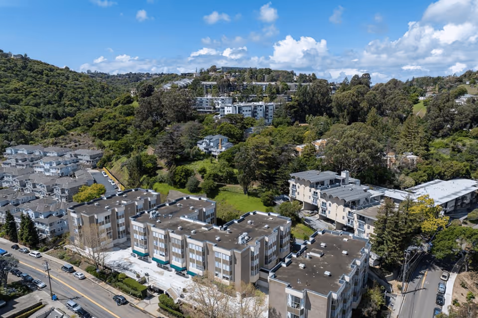 Aerial view of a residential facility complex named Bonnie Brae Terrace surrounded by lush green hills and trees under a partly cloudy blue sky.