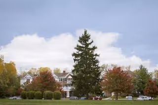 Exterior view of a multi-story senior living facility building surrounded by trees with autumn foliage and a large evergreen tree in the foreground under a partly cloudy sky.