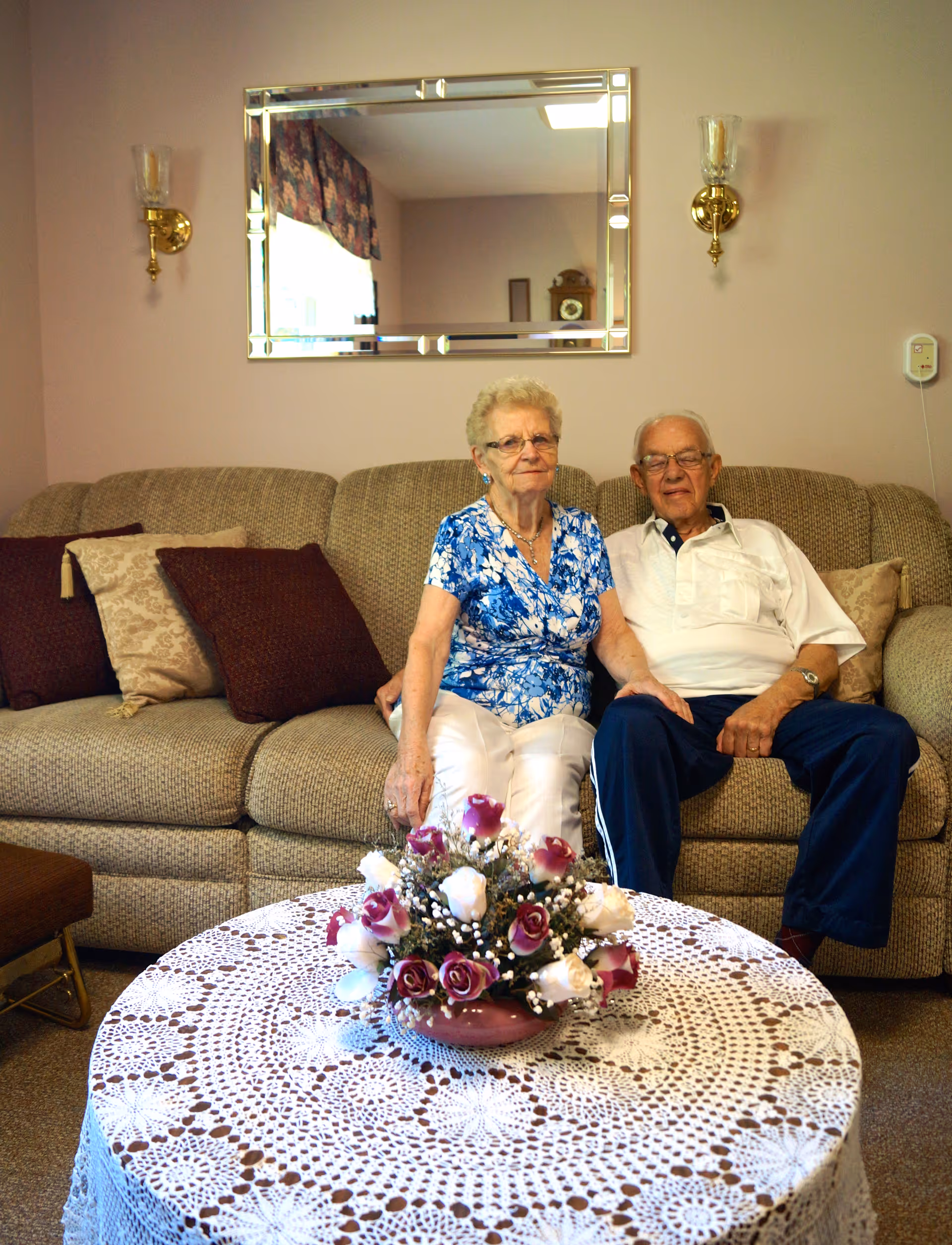 An elderly couple sitting on a sofa in a living room with a lace-covered coffee table and a floral centerpiece.