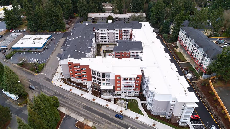 Aerial view of a large multi-story senior living facility named The Reserve At Lynnwood, surrounded by trees and adjacent buildings, with parking areas and roads visible.