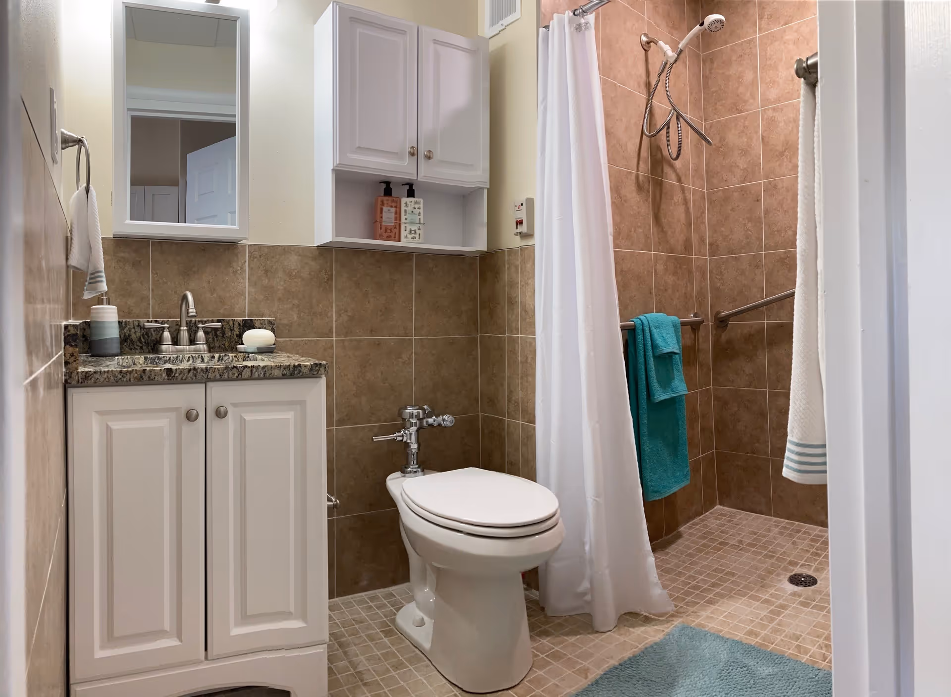 A clean bathroom featuring a white toilet, a vanity with a granite countertop and a sink, a mirror above the sink, and a shower area with brown tiled walls and a white shower curtain. There are teal and white towels hanging on grab bars inside the shower and on a towel ring beside the sink.
