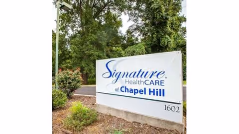 Outdoor view of a white sign with blue text that reads 'Signature HealthCARE of Chapel Hill' located near a road with trees and bushes in the background.