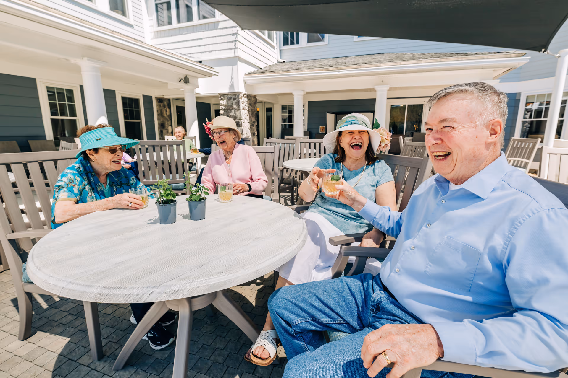 Four elderly people sitting around a round outdoor table at a senior living facility, enjoying drinks and laughing together on a sunny day. The setting includes patio furniture, potted plants on the table, and a building with white columns and blue siding in the background.