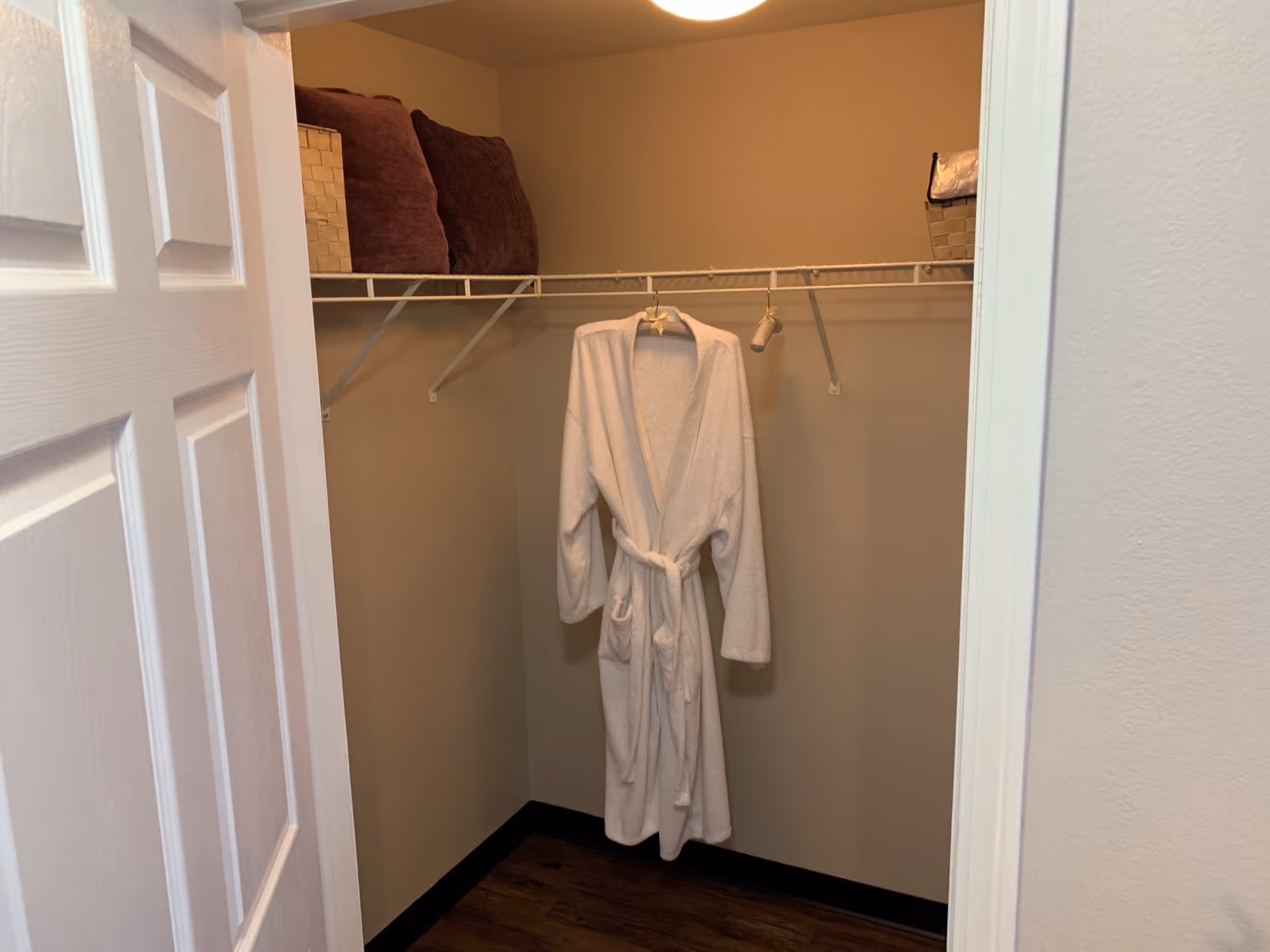 A small walk-in closet with a white bathrobe hanging on a wire shelf. The shelf also holds two brown cushions and a basket with folded linens. The closet has beige walls and a wooden floor.