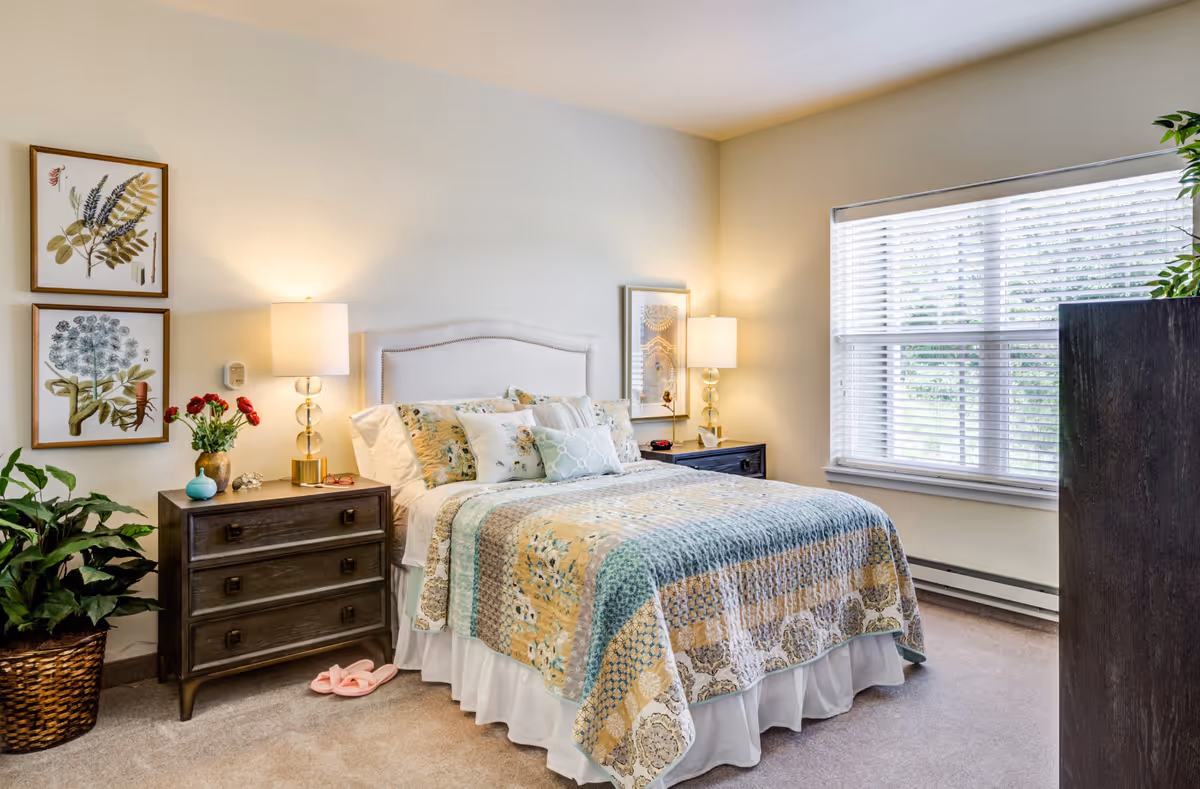 Well-lit bedroom with a patterned quilted bed, two nightstands with lamps, framed botanical art, a potted plant, and a large window with blinds.