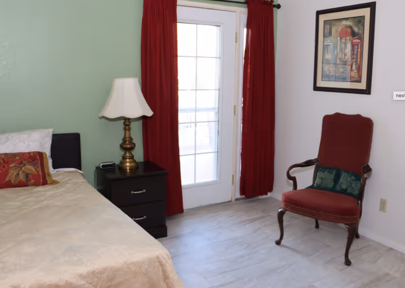 Sunlit bedroom with a bed, nightstand and lamp, red curtains over a glass door, and a red upholstered chair.
