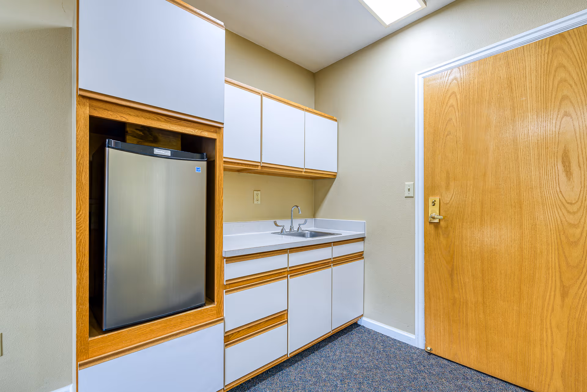 Small kitchenette area with a stainless steel mini fridge, white cabinets, a sink and a wooden door.