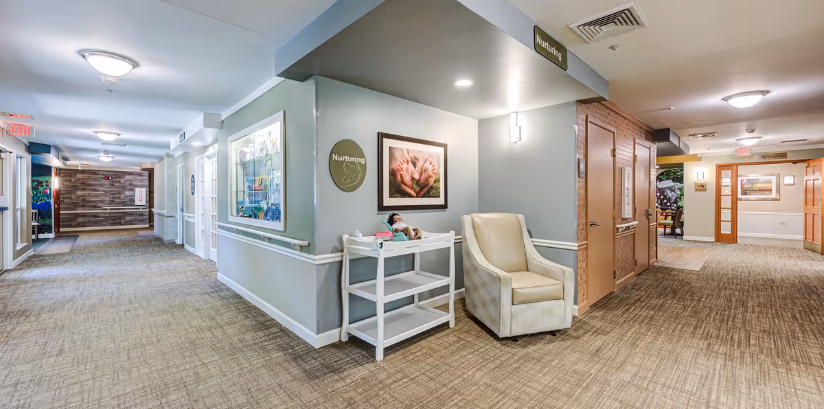 A well-lit hallway in a senior living facility with beige carpet and light blue walls. On the right side, there is a beige armchair and a white changing table with a doll on it beneath a framed picture of adult hands holding baby feet. Signs labeled 'Nurturing' are visible on the wall and ceiling. The hallway extends in two directions with doors and handrails along the walls, and exit signs are visible in the distance.