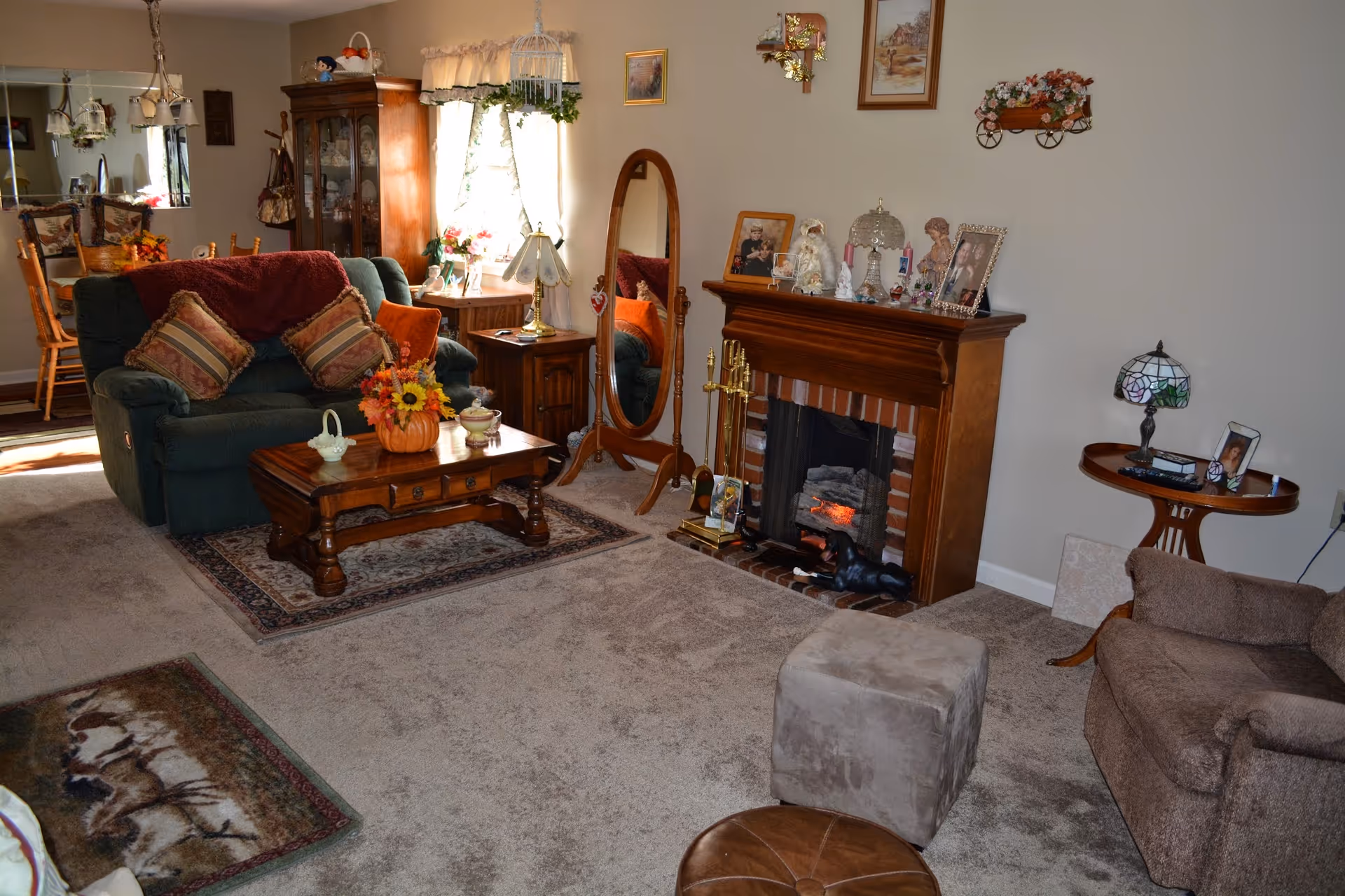 A cozy living room with a green loveseat adorned with patterned pillows and a red throw blanket, a wooden coffee table with a pumpkin-shaped flower arrangement, a fireplace with decorative items on the mantel, a standing oval mirror, a small side table with a lamp and framed photo, and a brown armchair with a matching ottoman. In the background, a dining area with wooden chairs and a china cabinet is visible.