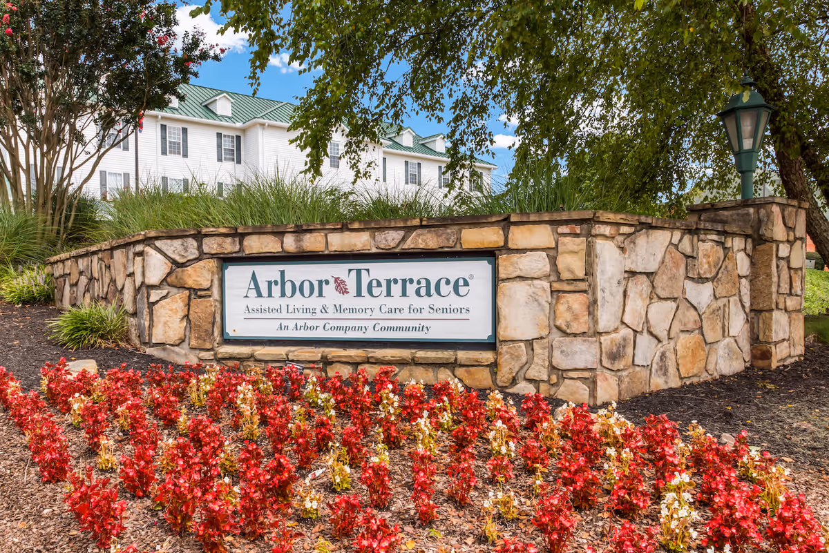 Stone sign for Arbor Terrace Assisted Living & Memory Care for Seniors surrounded by red flowers and greenery, with a white building and green roof in the background under a blue sky with clouds.