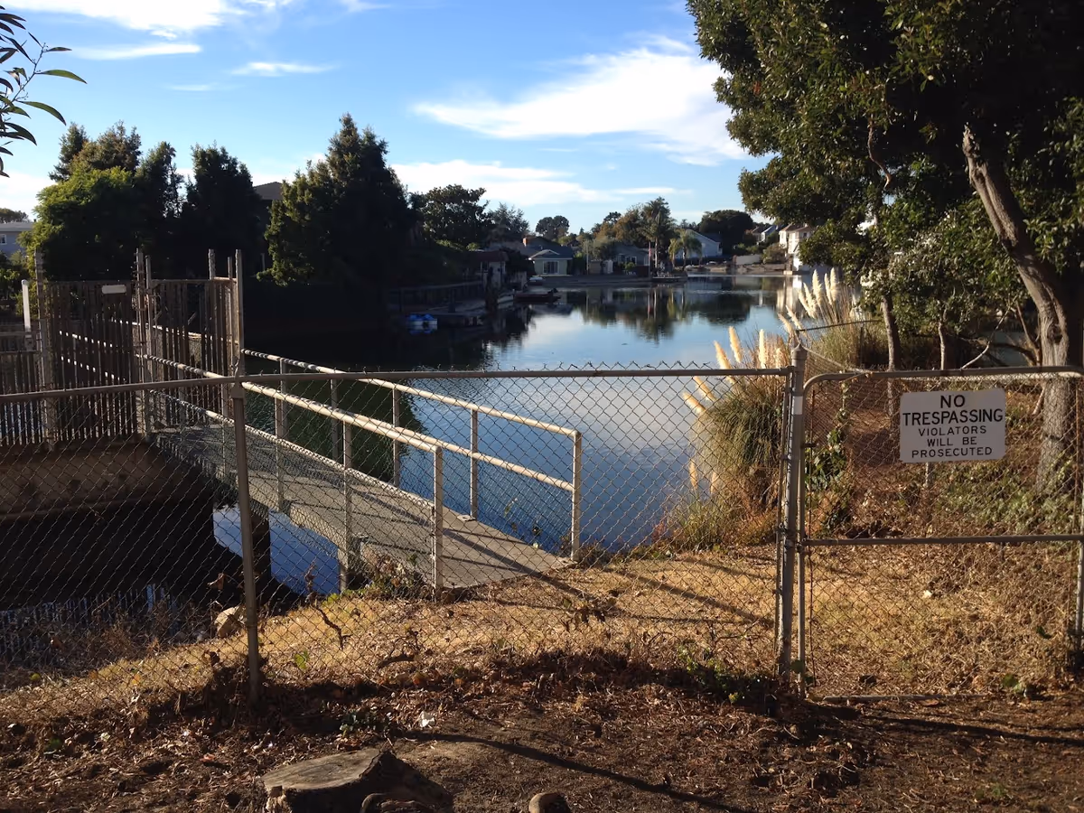 A fenced area with a chain-link gate and a 'No Trespassing' sign overlooking a calm water body surrounded by trees and houses under a blue sky with some clouds.