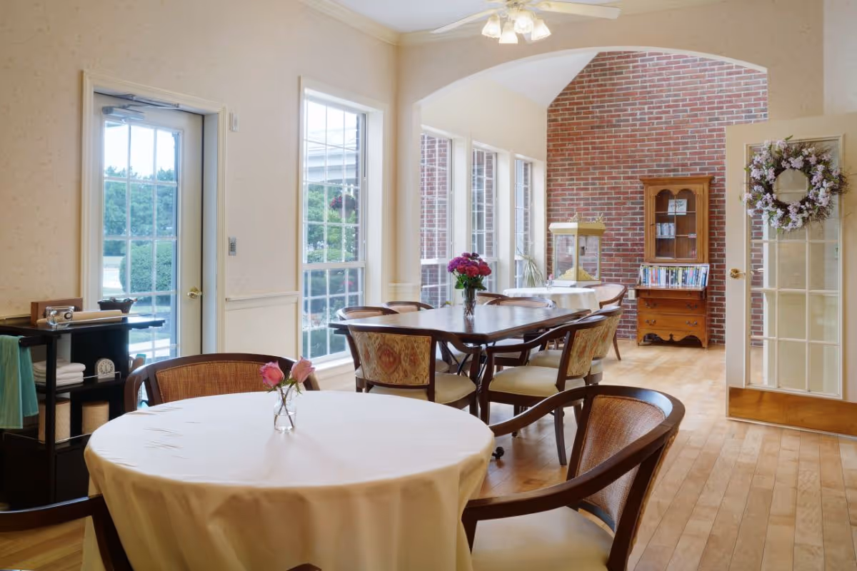 Sunlit communal dining room with round and rectangular tables and chairs, floral centerpieces, large windows, and a brick accent wall.