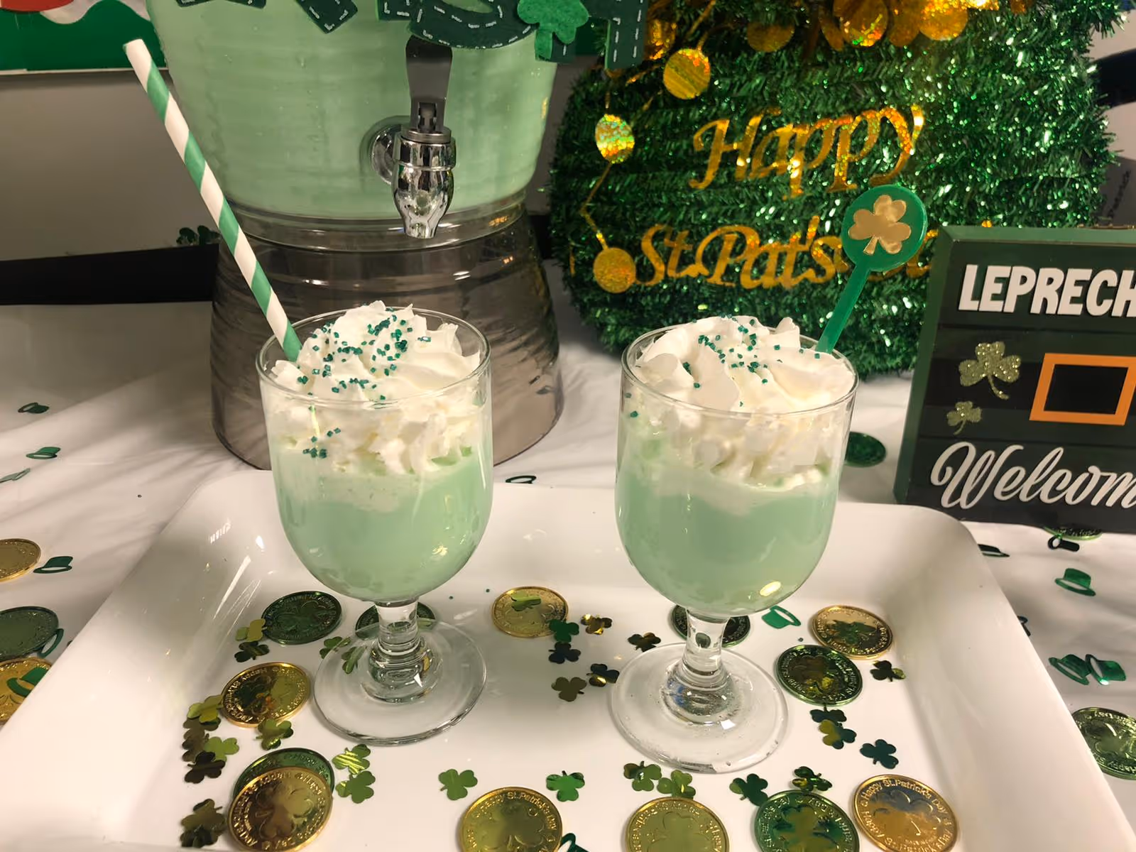 Two glasses filled with a green beverage topped with whipped cream and green sprinkles, each with a straw or stirrer, placed on a white tray decorated with gold and green shamrock confetti and coins. In the background, there is a green beverage dispenser and a green glittery decoration with the words 'Happy St. Pat's'. A sign reading 'LEPRECHAUN Welcome' is also visible.
