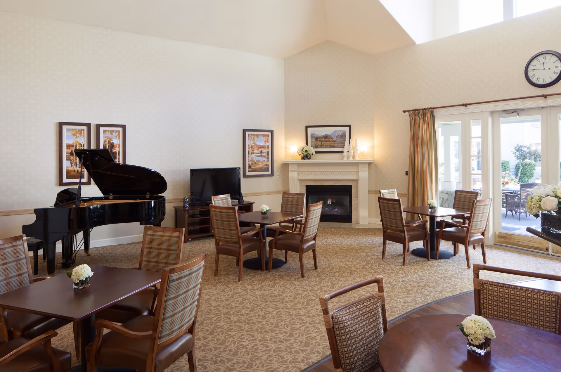 Bright senior living common room with several tables and chairs, a grand piano, fireplace, TV and glass doors leading to an outdoor patio.