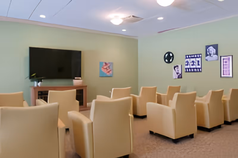 A small theater-style room with beige armchairs arranged in rows facing a large flat-screen TV mounted on a light green wall. The room has ceiling lights and several framed pictures and film-themed decorations on the walls.