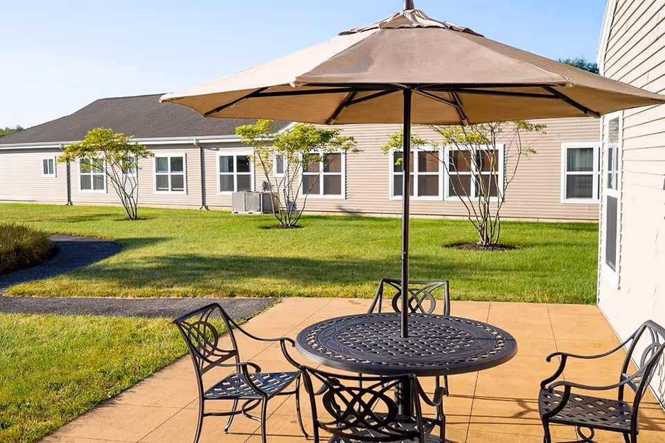 Outdoor patio area with a round metal table and four matching chairs under a large beige umbrella. The patio is adjacent to a beige building with multiple windows, surrounded by green grass and small trees.