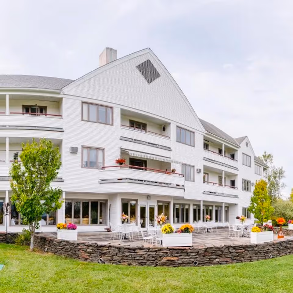 Exterior view of a multi-story white building with balconies and large windows. The building has a stone patio area with white metal tables and chairs, decorated with colorful flower arrangements. There is green grass and trees surrounding the patio.