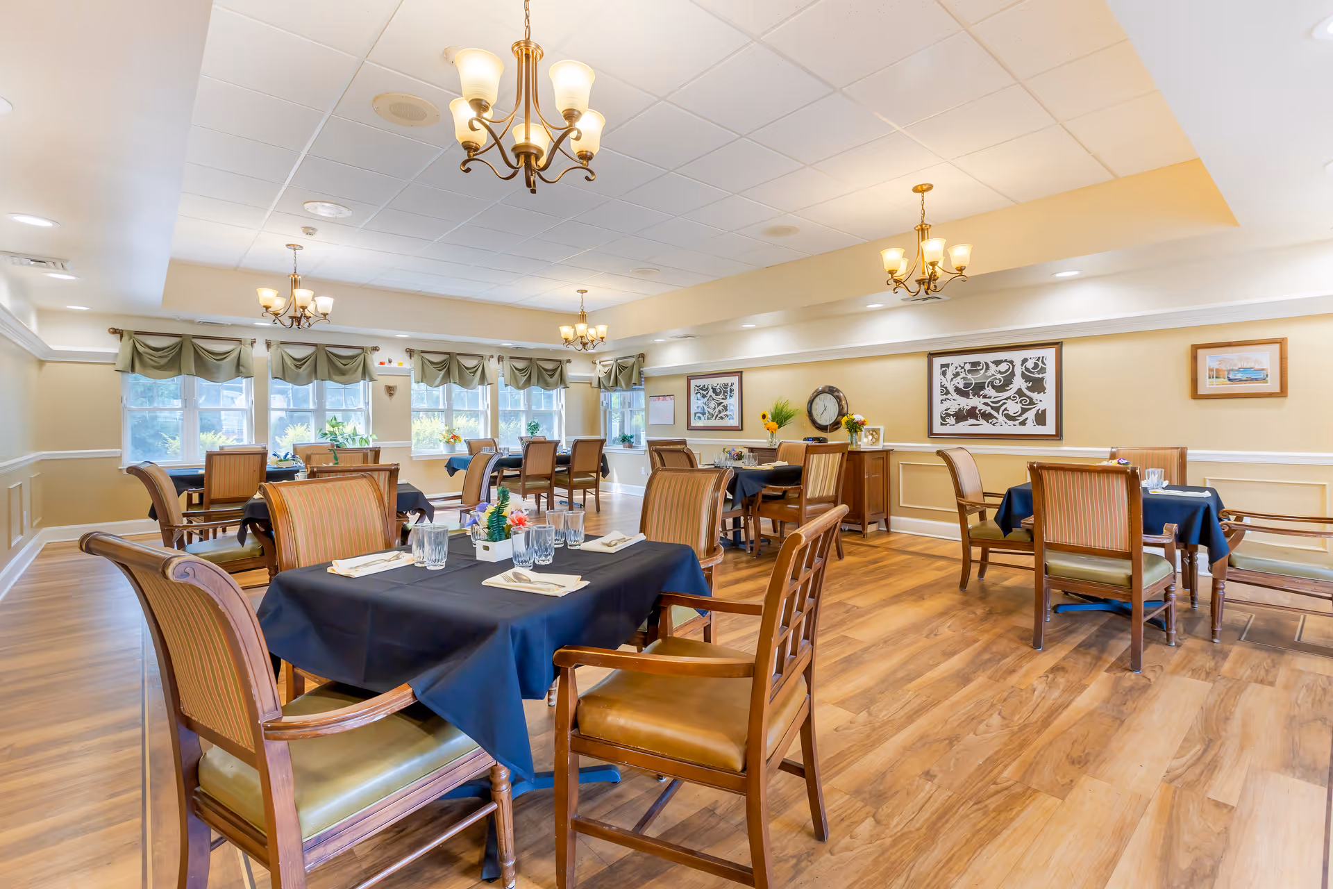 Well-lit dining room with multiple tables covered in dark tablecloths, wooden chairs, chandeliers, and windows letting in natural light.
