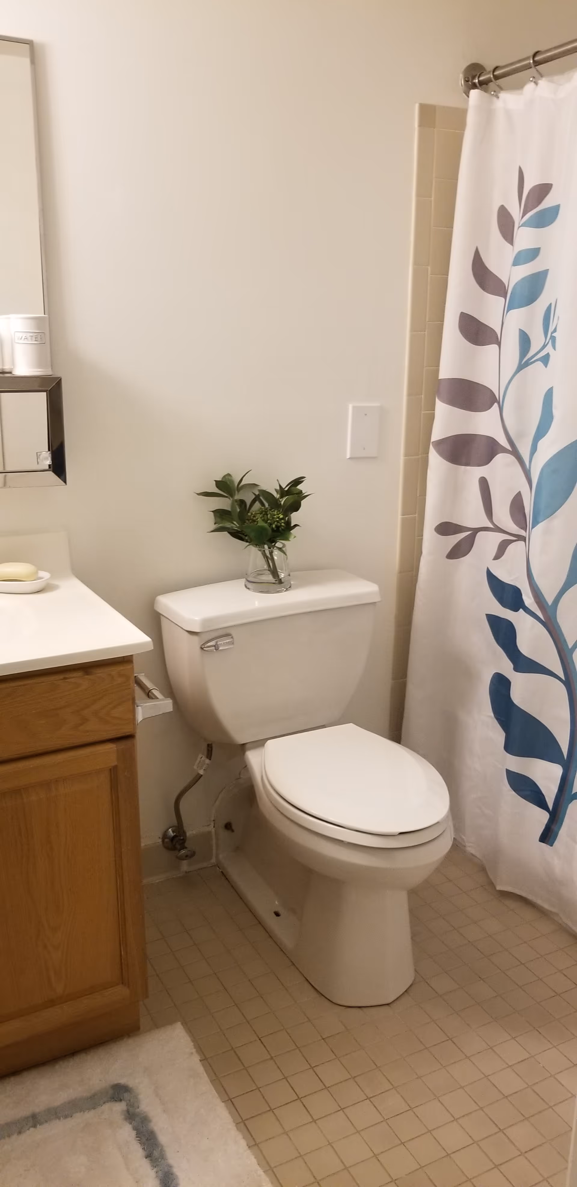 A clean bathroom featuring a white toilet with a small glass vase holding green leafy branches placed on top of the tank. To the left is a wooden vanity with a white countertop, a soap dish with soap, and a mirrored medicine cabinet above. To the right is a shower area with a white curtain decorated with blue and purple leaf patterns. The floor is covered with beige square tiles and a white bath mat with a blue border is partially visible.